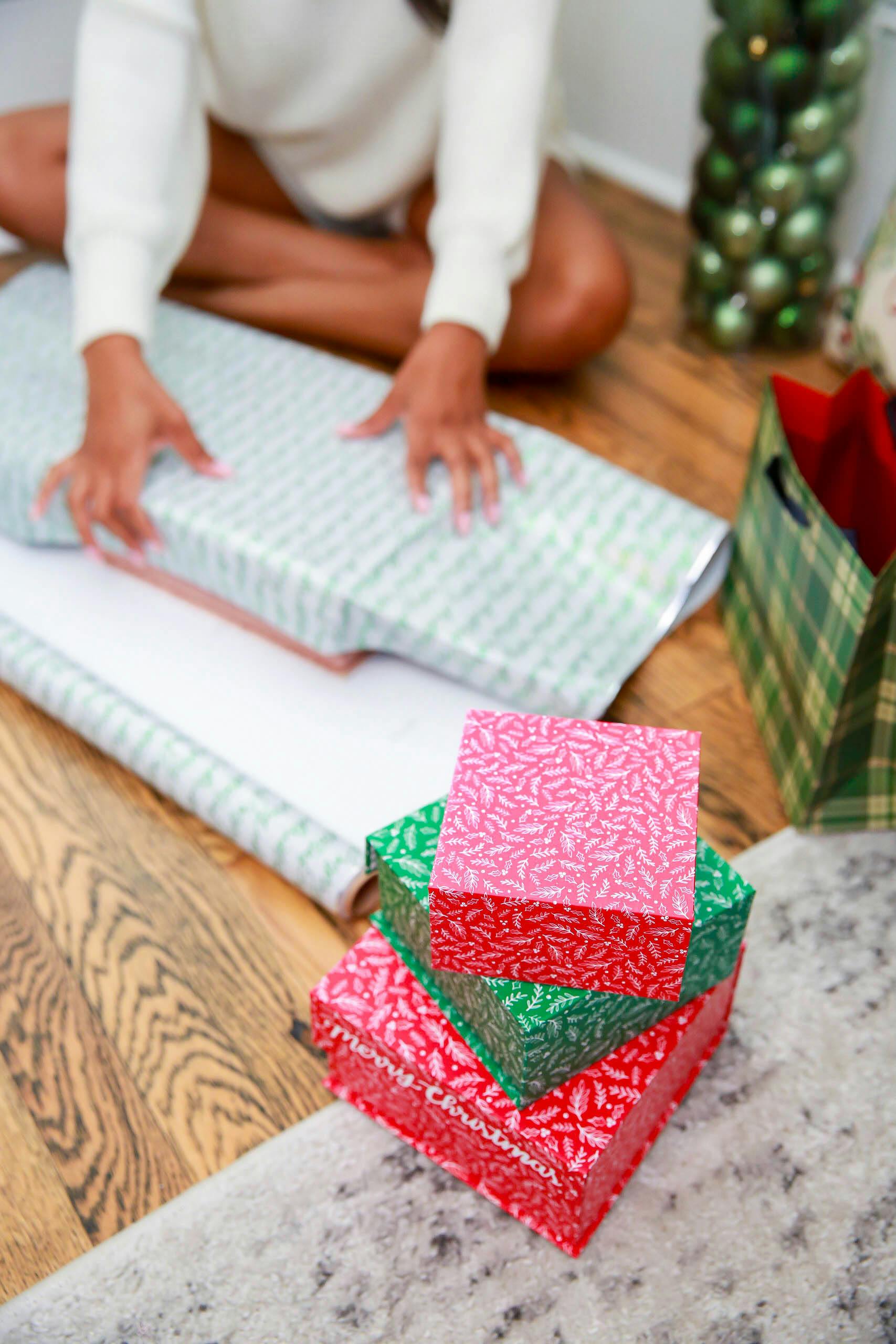 Woman wrapping Christmas gifts on wood floor, surrounded by festive decor and colorful stacks.